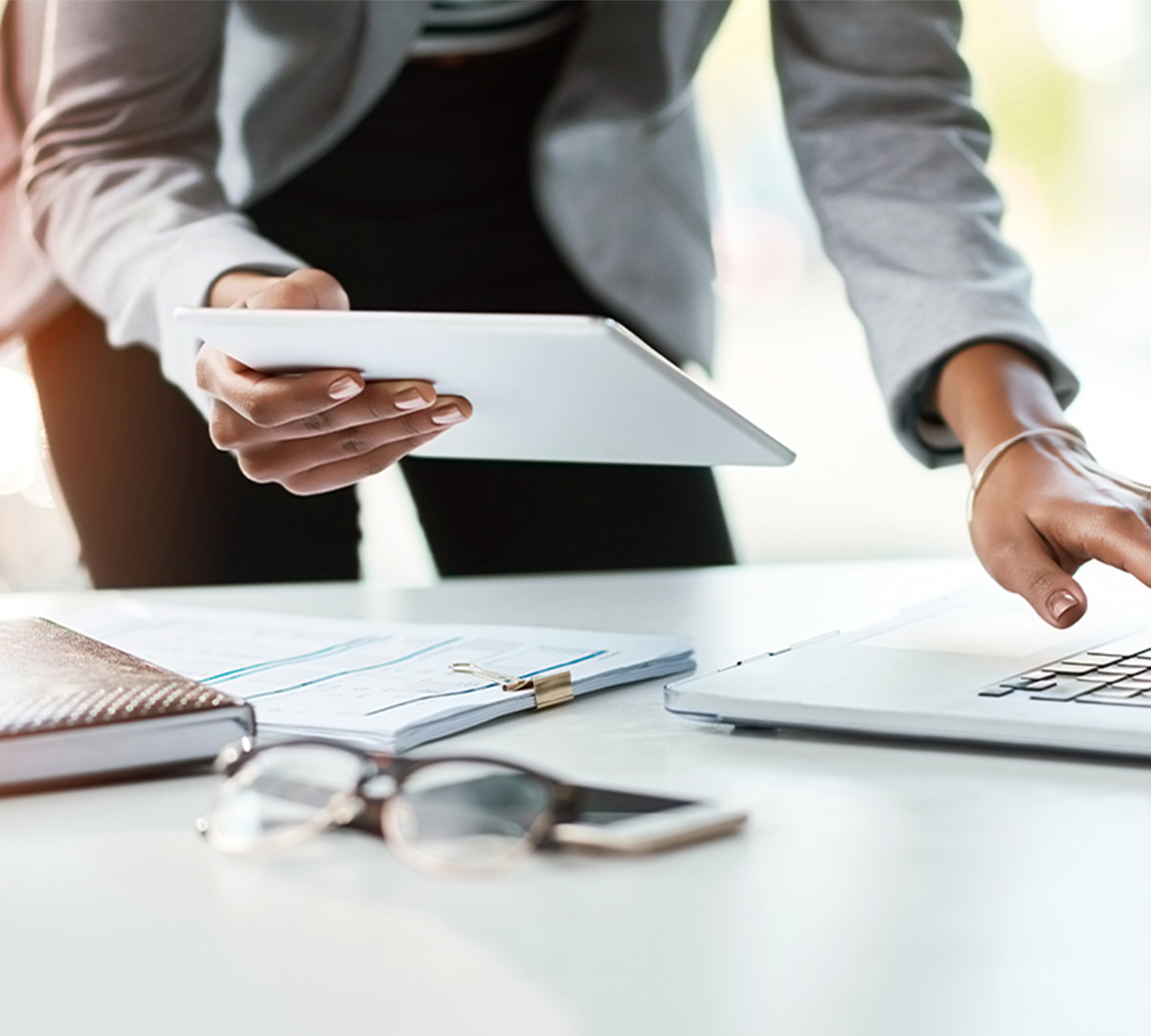 person standing over desk holding a tablet and working on a laptop