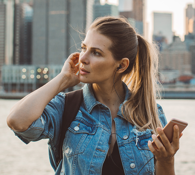Woman using ear buds to listen to news with her mobile. New York City in the background.