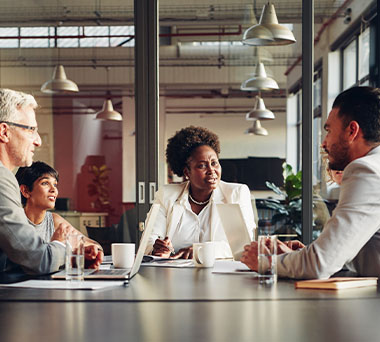 Three people at a conference room table discussing over laptops and paper