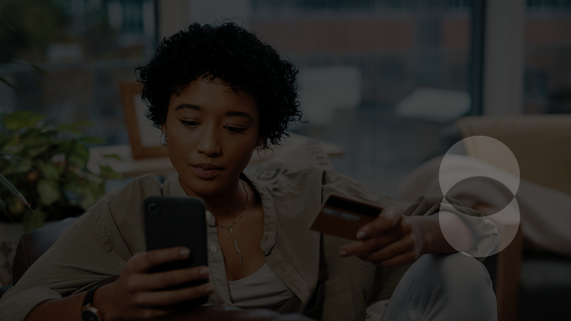Person on sofa preparing to buy something on their mobile phone while holding a credit card in the other hand.
