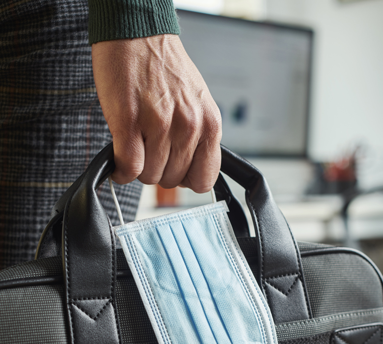 close up of hand holding face mask and laptop bag in office setting