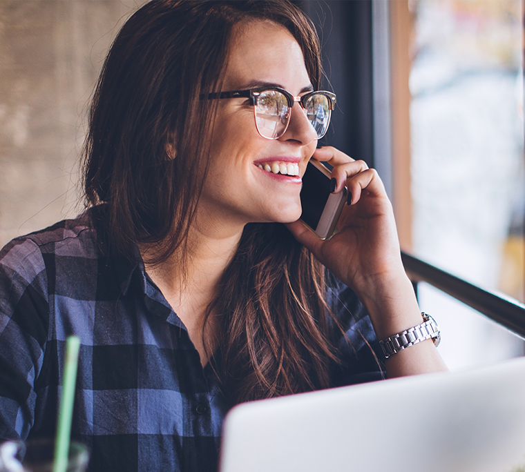 Person on phone, sitting in front of a window in a café with a laptop open on the table in front of them.